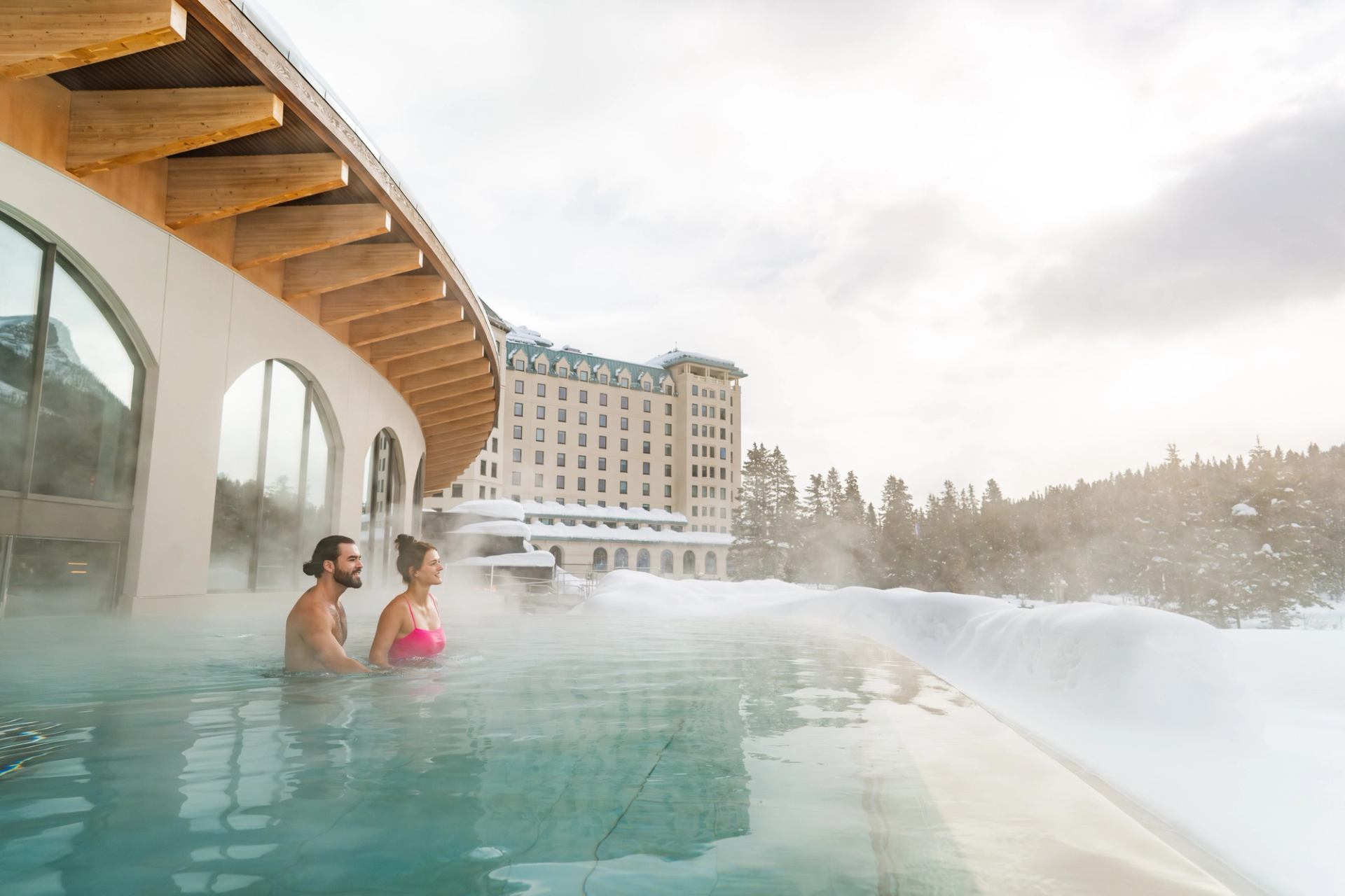 A couple in an outdoor pool look to the mountains on a snowy day.