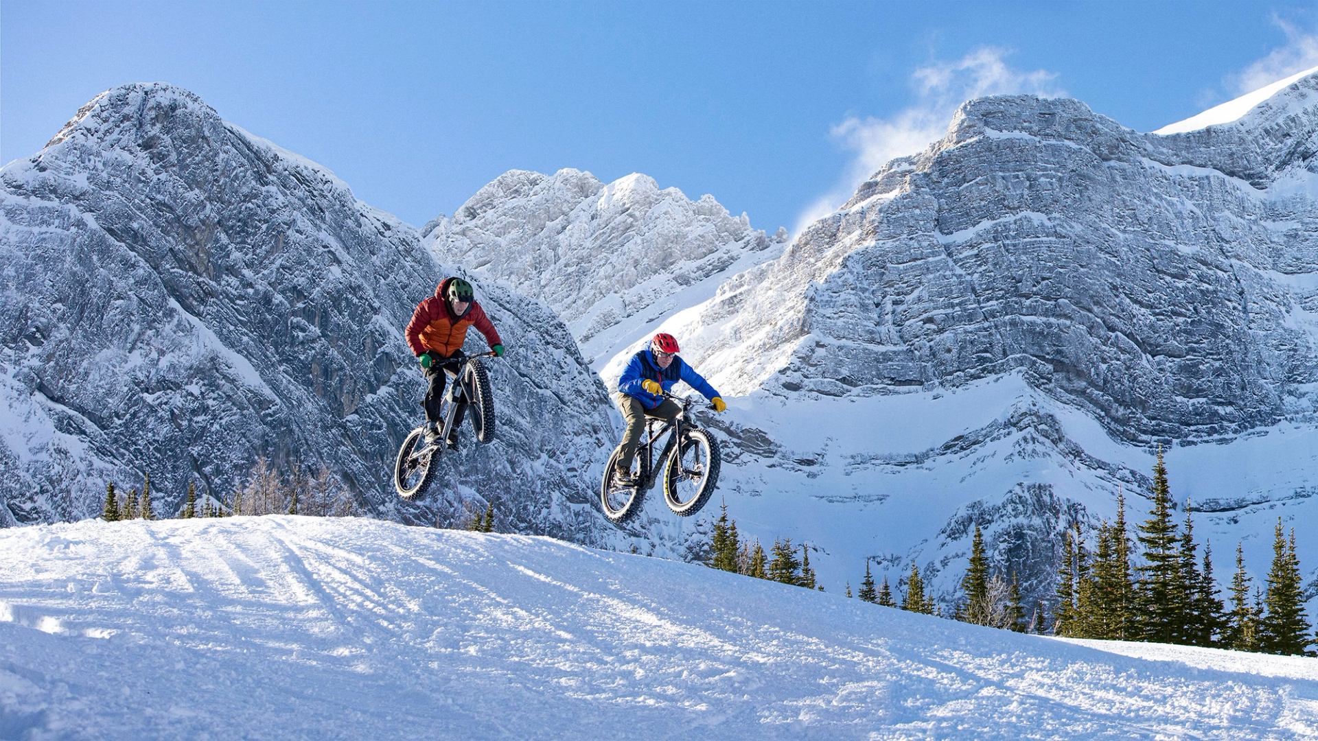 A panoramic shot of two people fatbiking in the winter with mountains in the background in Kananaskis Country.