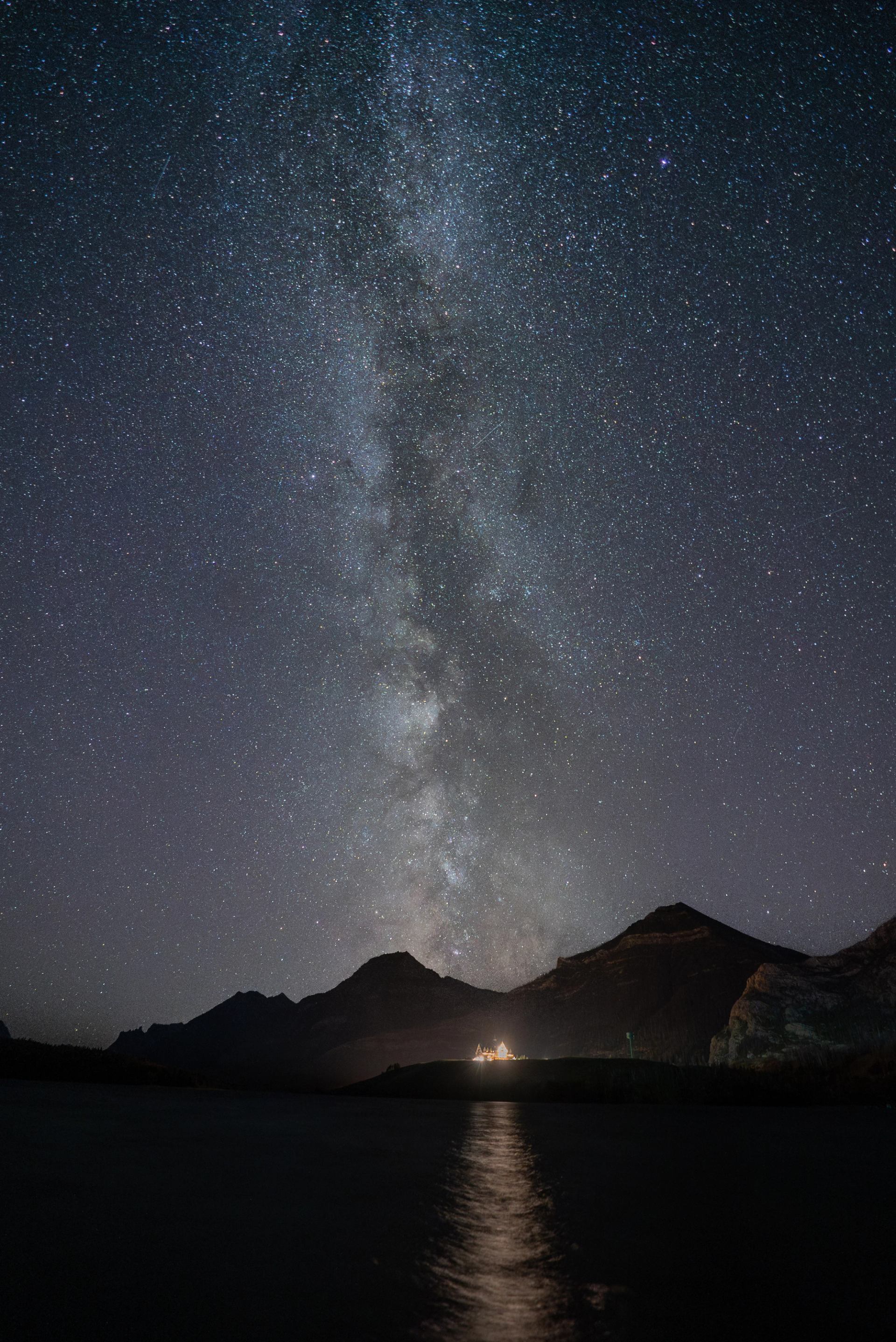 The Milky Way and countless stars glow above mountains and the brightly lit Prince of Wales Hotel.