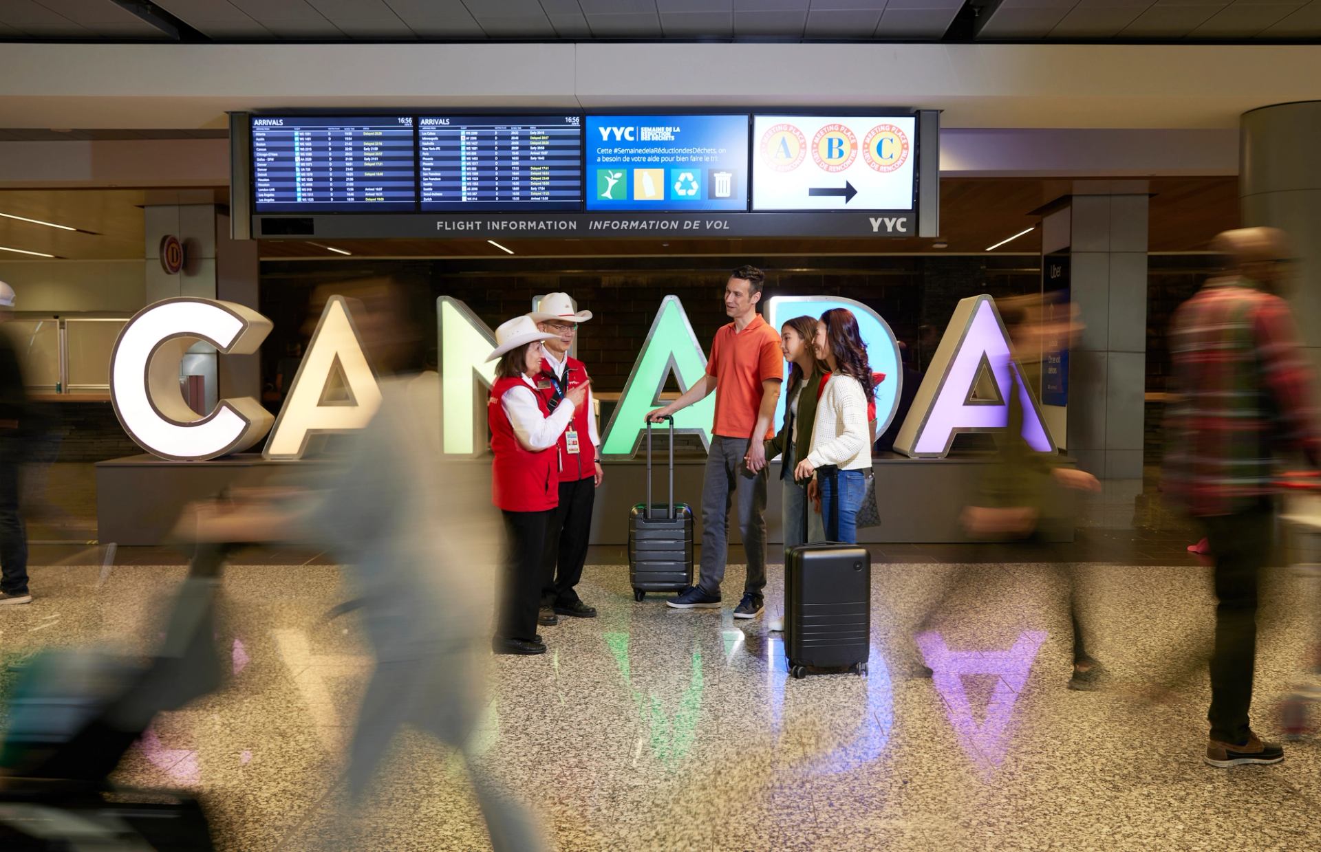 YYC Calgary International Airport white hat volunteer greeters talk with a family near a Canada sign.