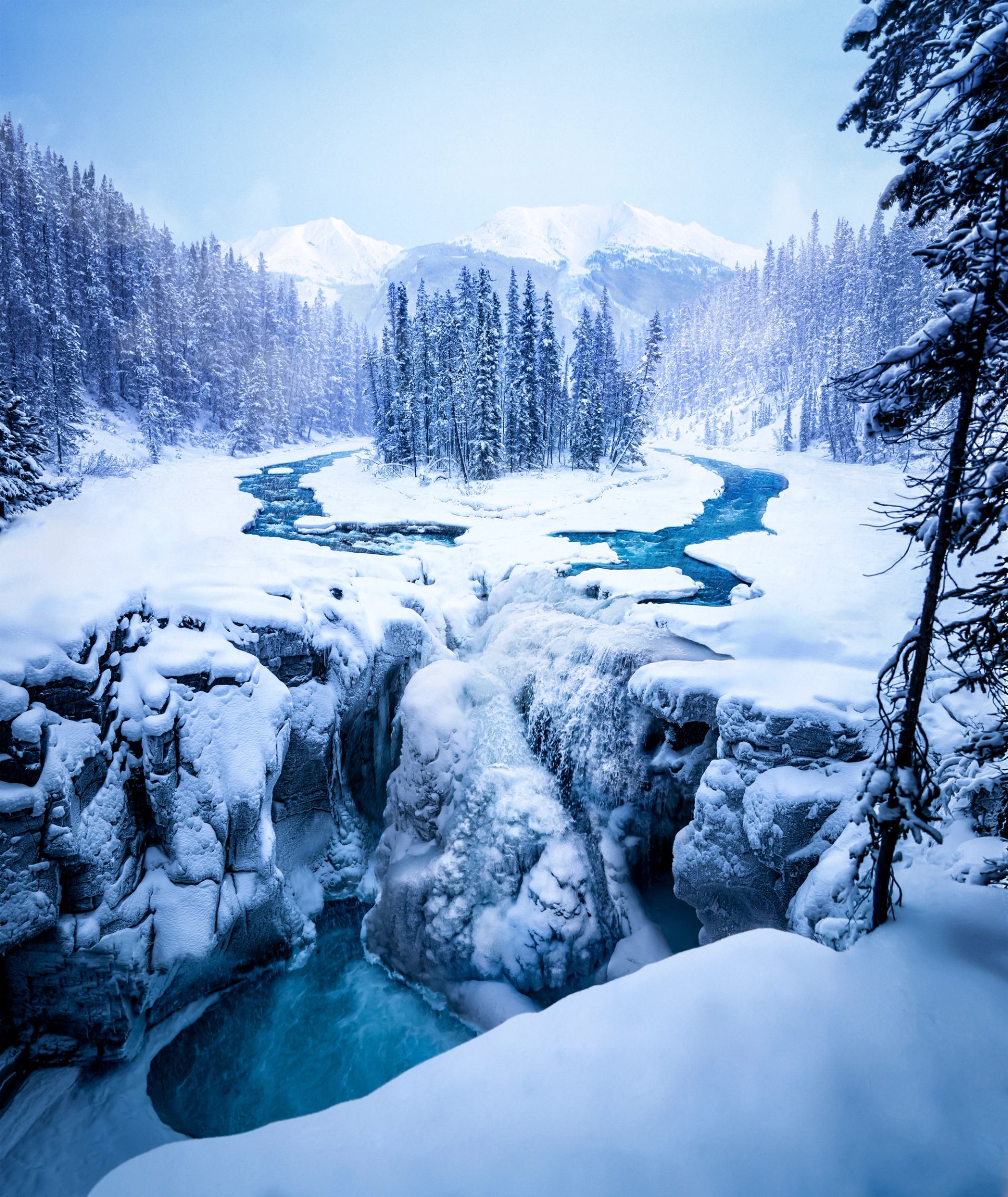 A frozen forest surround a small island in a river that splashes into a half-frozen waterfall.