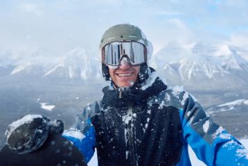 A man standing with his snowboard on a mountain at Lake Louise Ski Resort, smiling with the Canadian Rockies in the background.