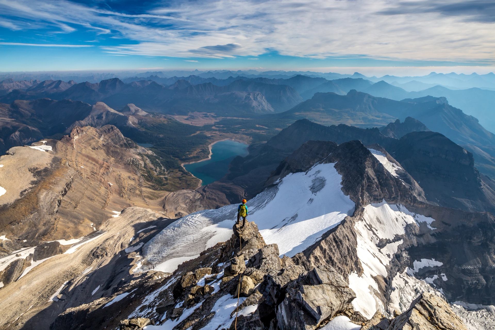 Wide shot of a hiker at the top of a lightly snow covered mountain peak over looking the expansive mountain range view