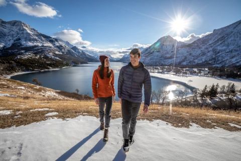 A couple on a winter hike, water, mountains and sunshine in background, in Waterton Lakes National Park.