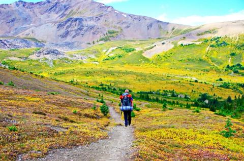 Backpacker with gear hiking on a trail toward the mountains in the background.