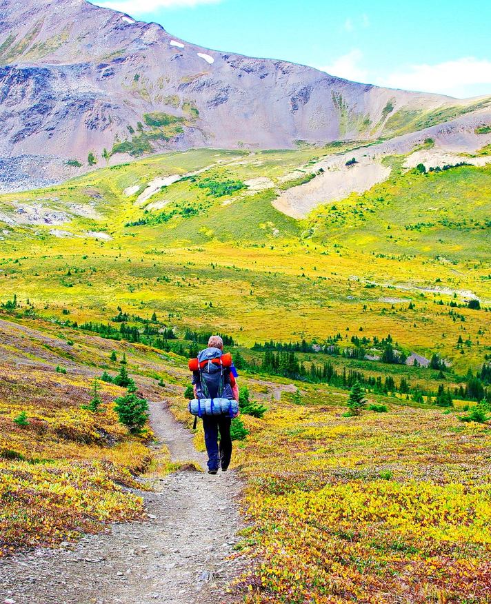 Backpacker with gear hiking on a trail toward the mountains in the background.
