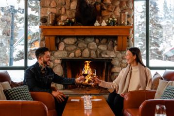 A man and woman smiling and tapping wine glasses while sitting on separate couches in front of a fireplace with a snow covered forest through the window in the background.