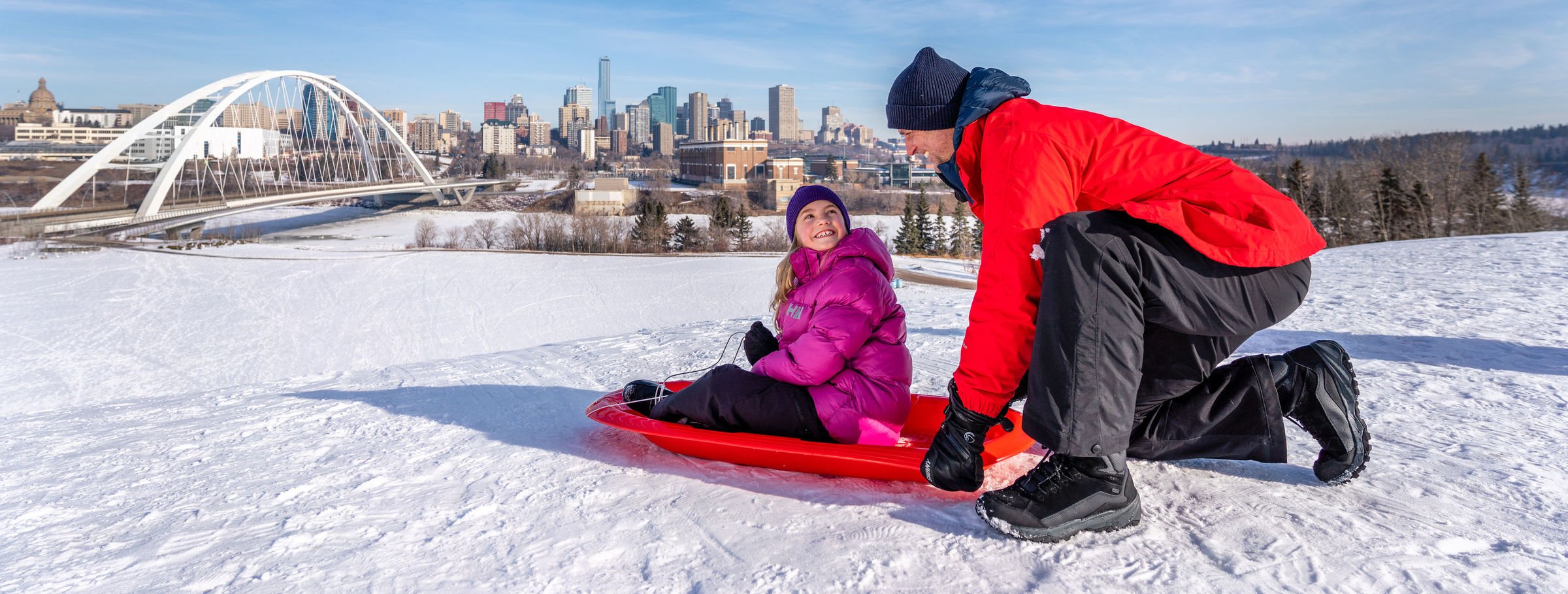 A father and daughter sledding with downtown Edmonton in the background.