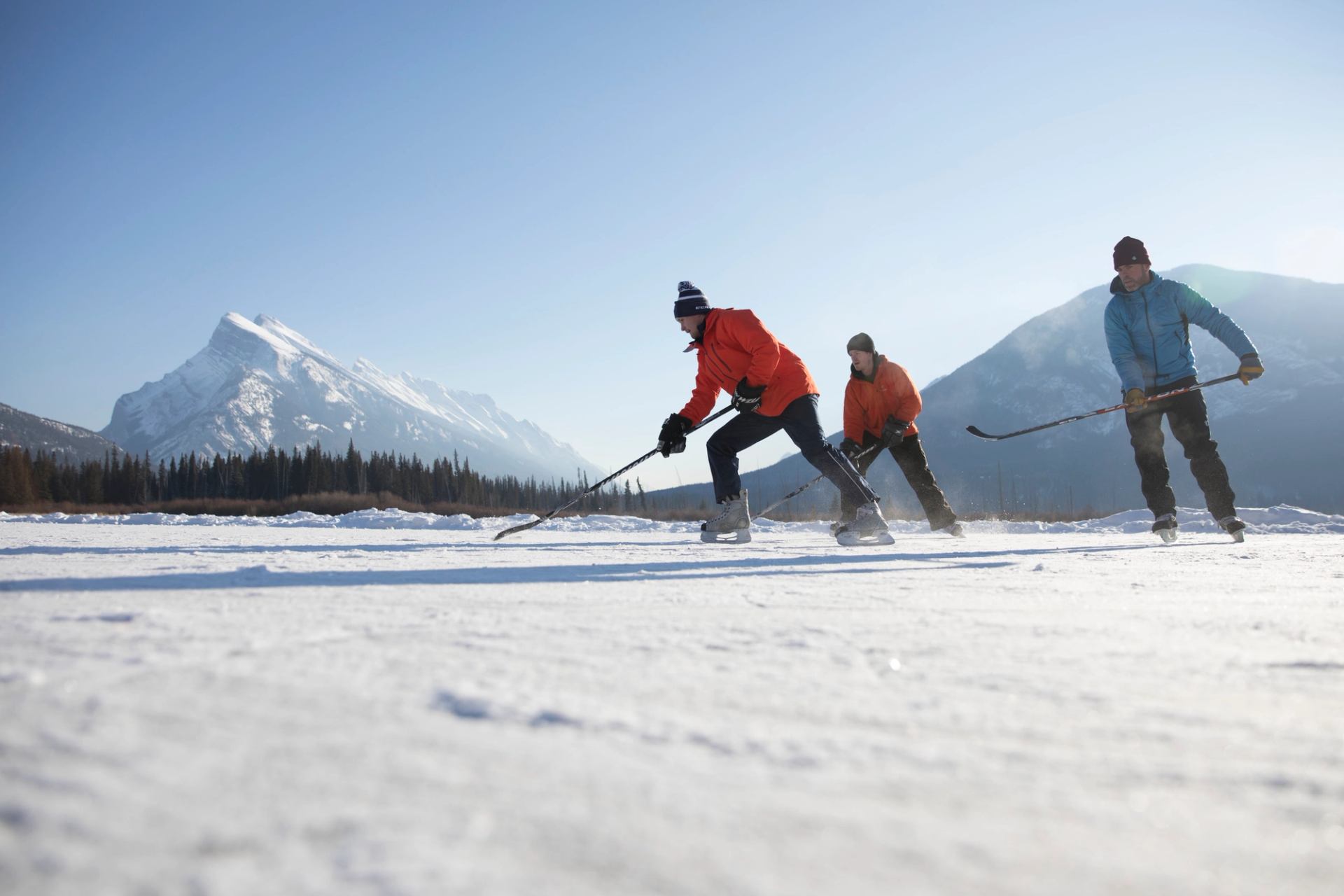 A group ice skating and playing pond hockey on a frozen lake with a mountain view.