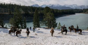 Actors on horseback surround Pedro Pascal's character Joel, on a field of snow near a river, with the mountains in the distance.