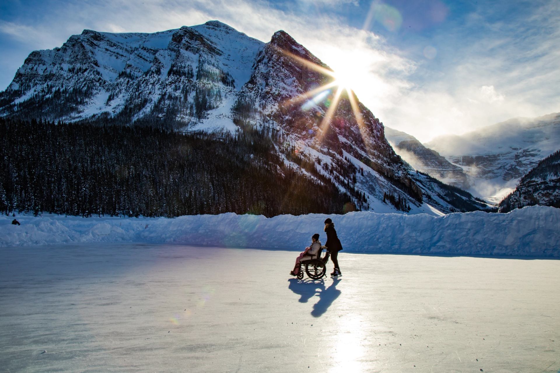 Son skating with his mom in a wheel chair with the sun peeking behind the mountains on Lake Louise in Banff National Park.