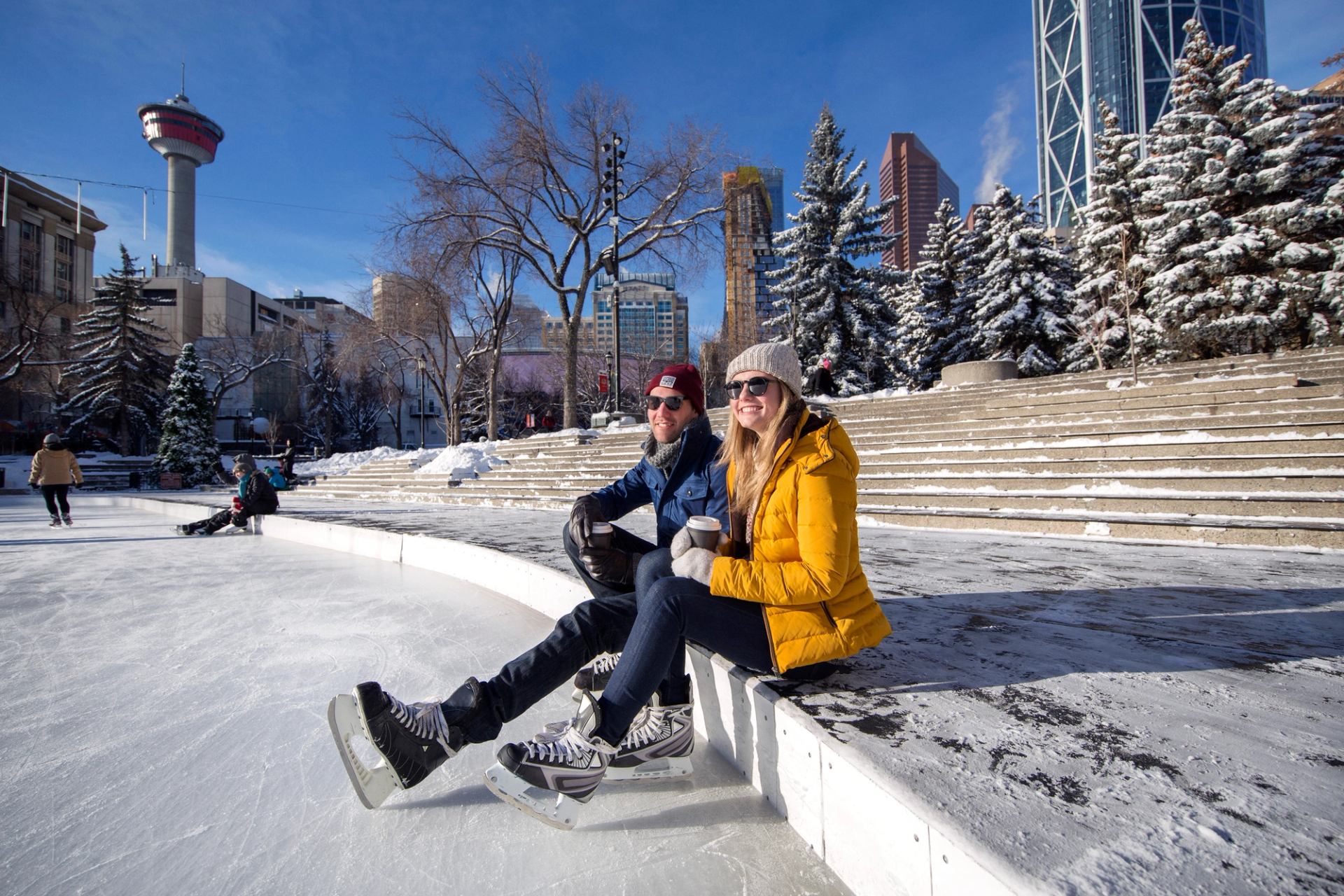 A couple sits on a ledge at an outdoor ice skating rink with  skates on holding a hot beverage with a downtown landscape in the background.