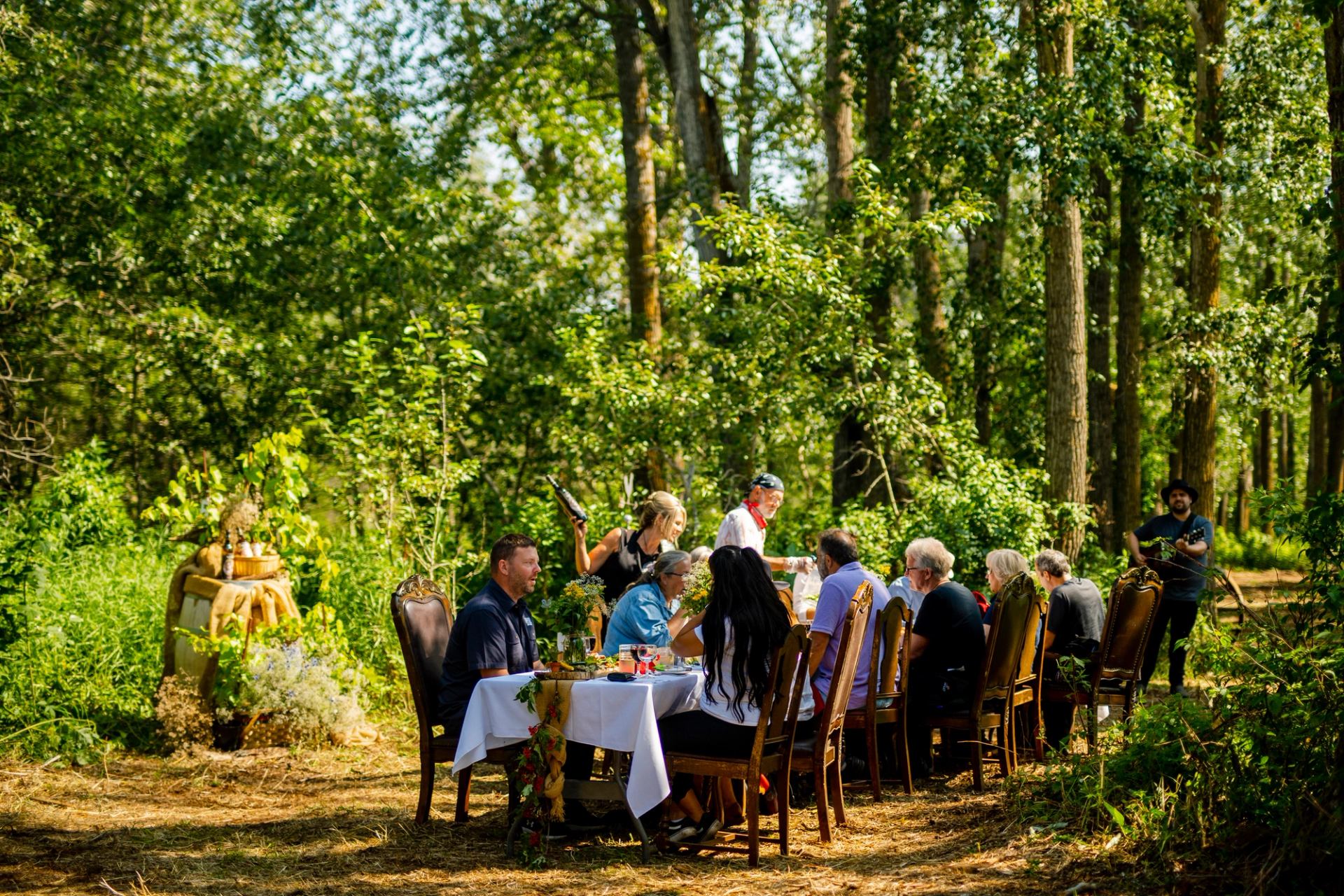 Group of people dining outdoors under a tree canopy at Prairie Gardens, whilst bathed in sunlight. Adventure Farm