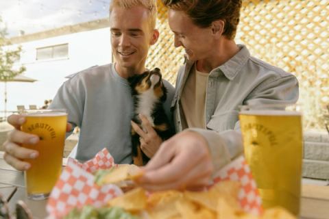 Two people with a dog have a beer and food at a restaurant.