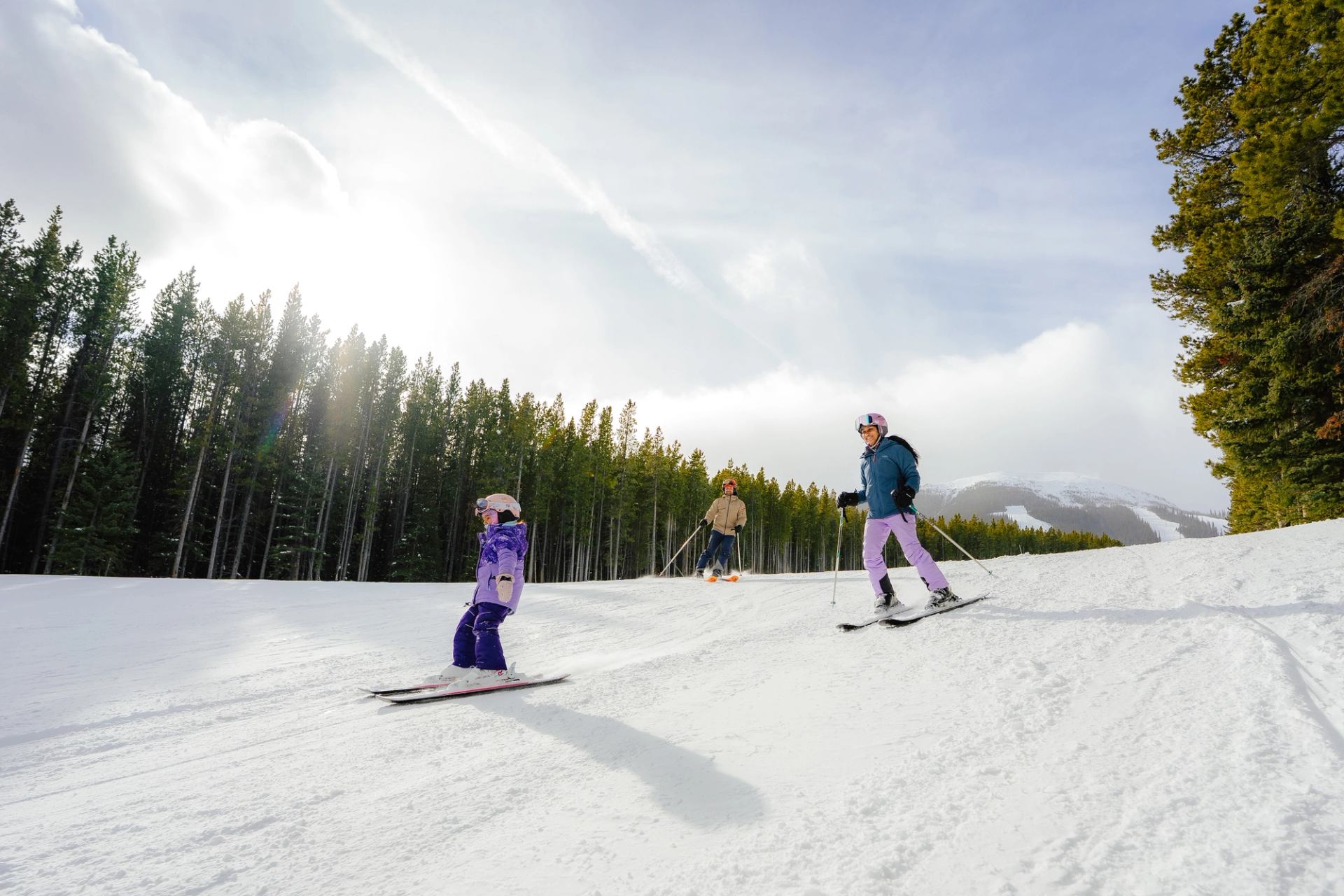 A family skis down a scenic trail at Nakiska with sunlight filtering through the trees