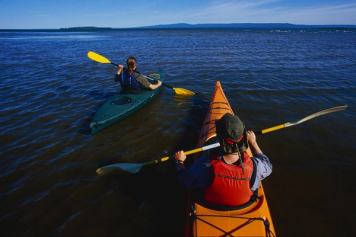 Two people paddle kayaks on a blue lake with a distant hill at shore.
