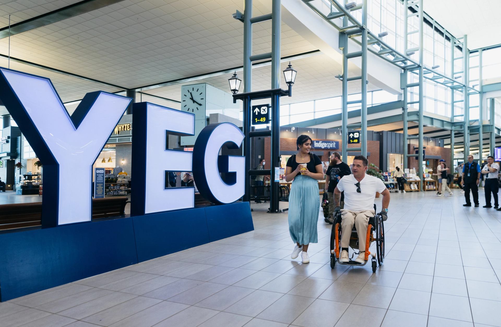 Woman and man in wheelchair pass the YEG letters sign at YEG Airport.