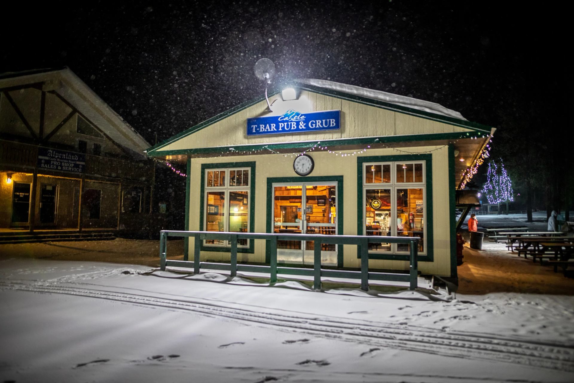 Warm light shines out the windows of the cozy, standalone T-Bar Pub & Grub at Castle Mountain Resort on a winter night while snow falls.