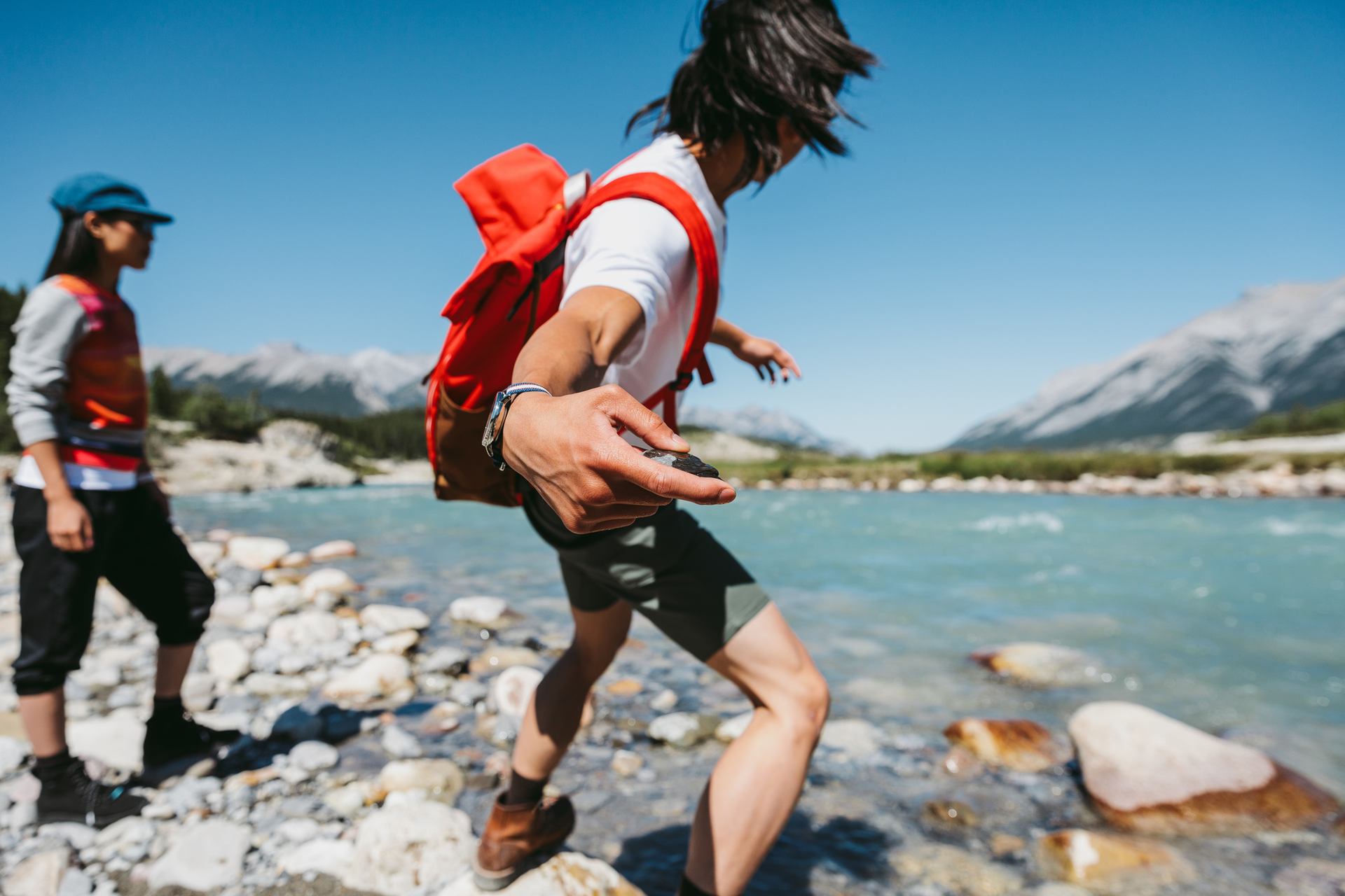 A man and woman skipping stones across a river with the mountains in the background.