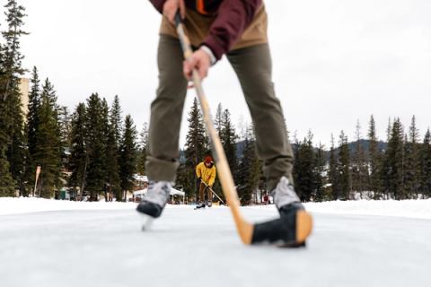 A ground-level shot of a puck and stick while friends play ice hockey on an outdoor rink.