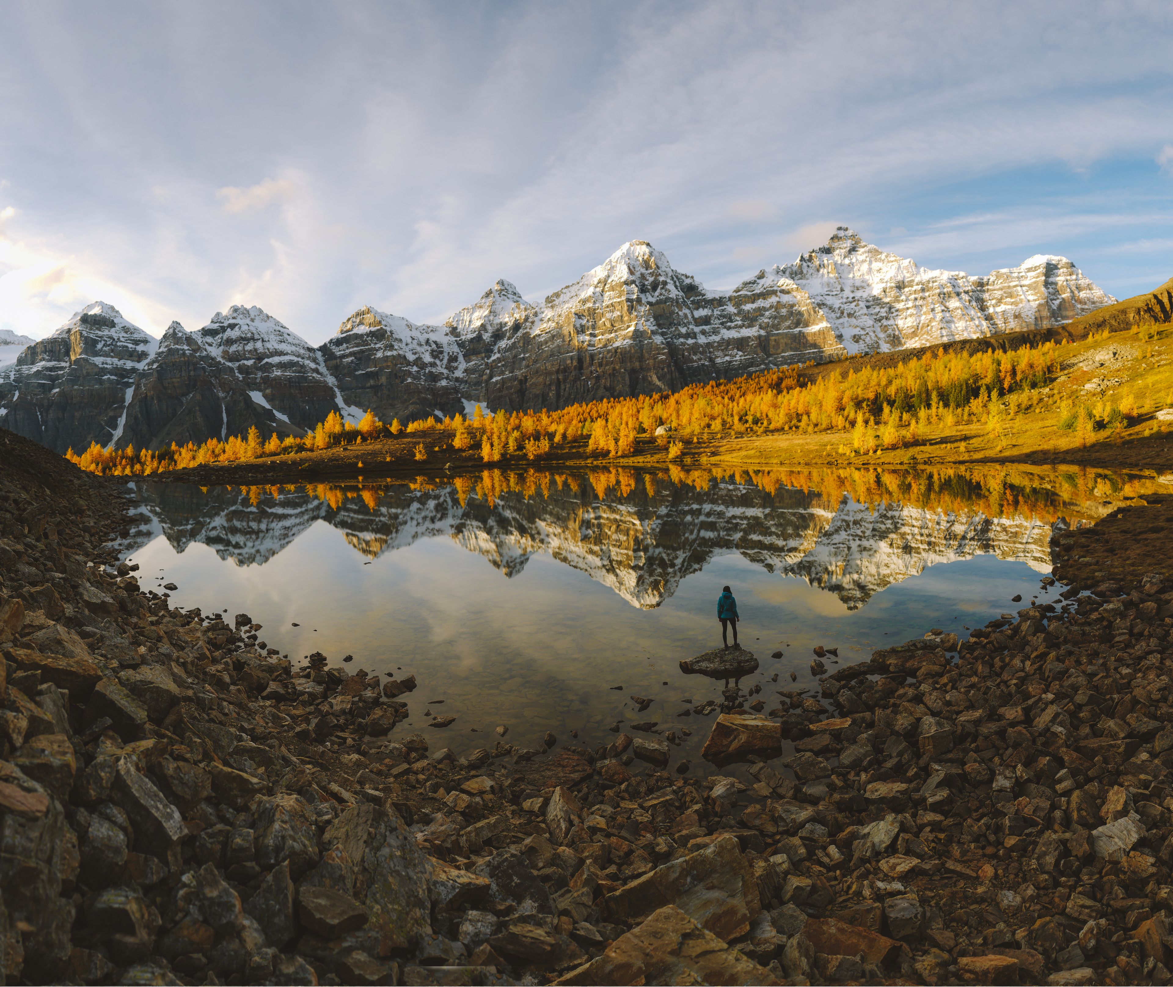 A hiker pauses at a small lake in Larch Valley, surrounded by golden trees and rocky mountains.