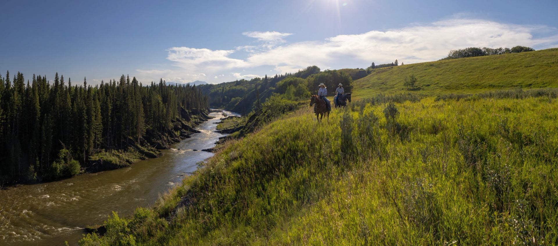 Two people riding horse alongside a riverbank