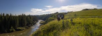Two people riding horse alongside a riverbank.