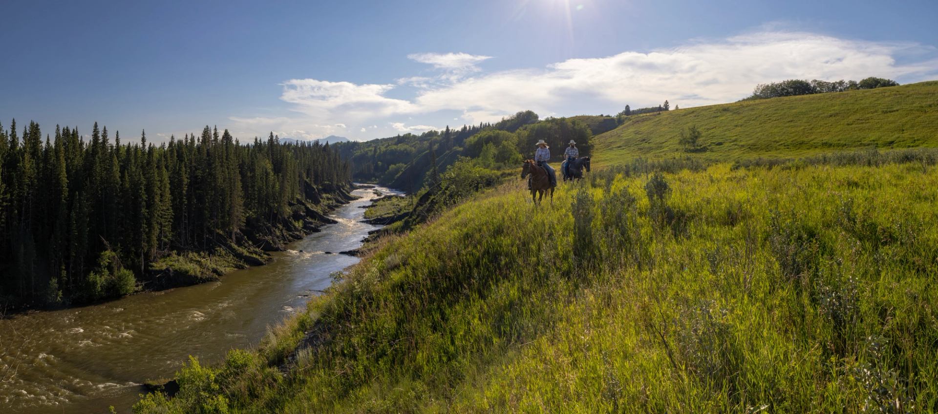 Two people riding horse alongside a riverbank