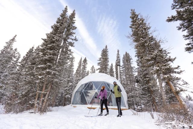 Couple cross-country skiing outside their glamping dome on a blue-sky winter day in Bragg Creek, Alberta
