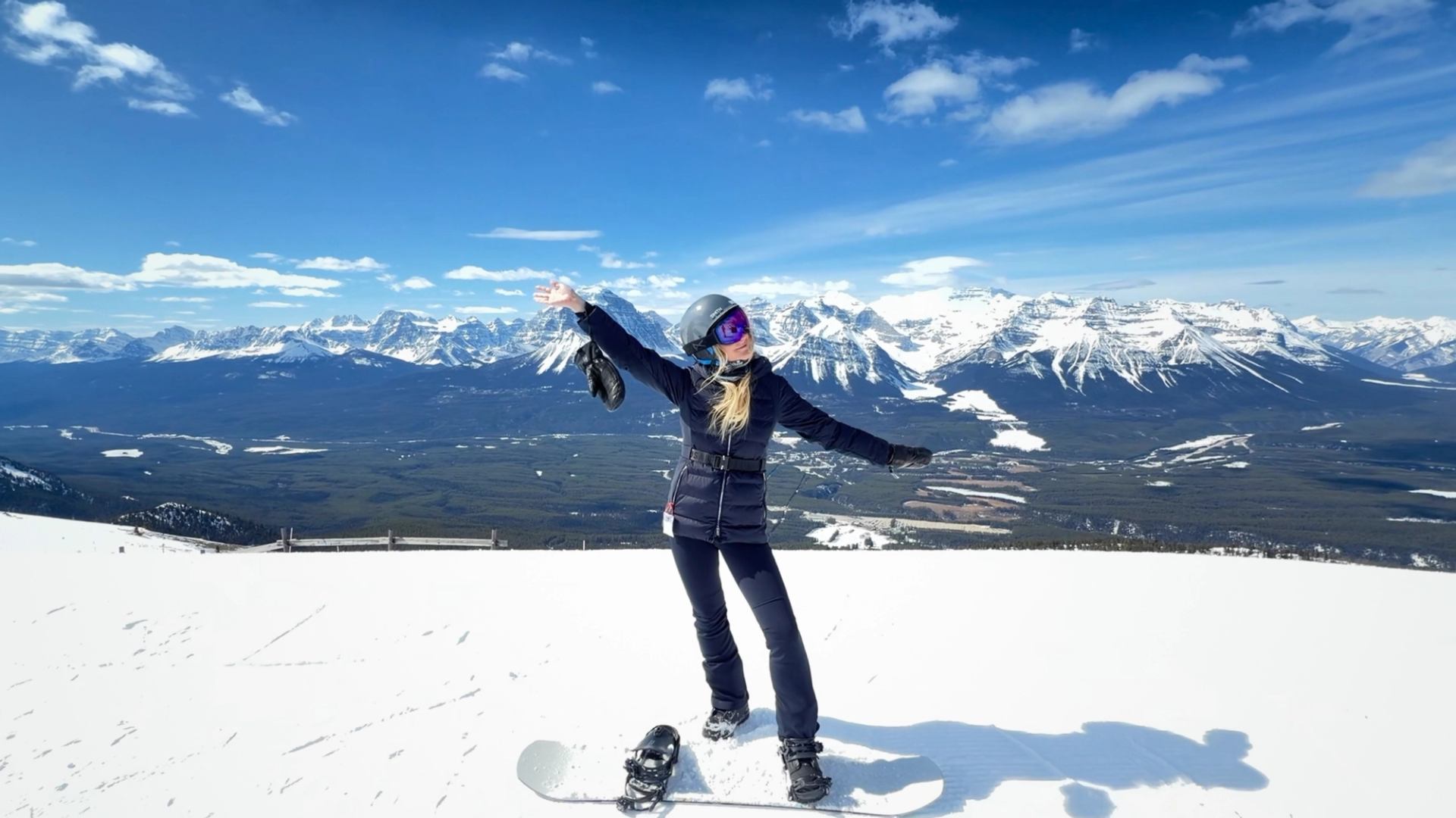 A snowboarder stands with her arms out at the top of a run at Lake Louise Ski Resort with mountains in the distance.