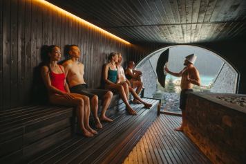 People relax in a luxurious sauna while a sauna master fans them, with a view of Lake Louise out a window.