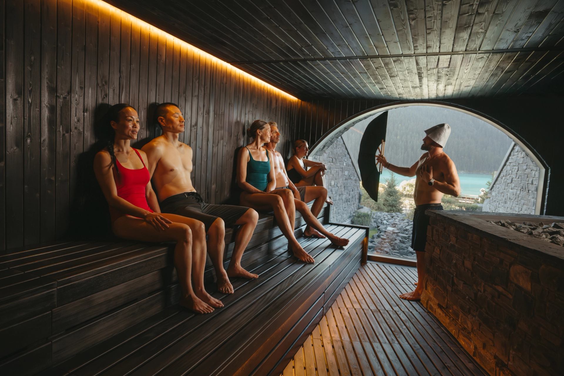 People relax in a luxurious sauna while a sauna master fans them, with a view of Lake Louise out a window.