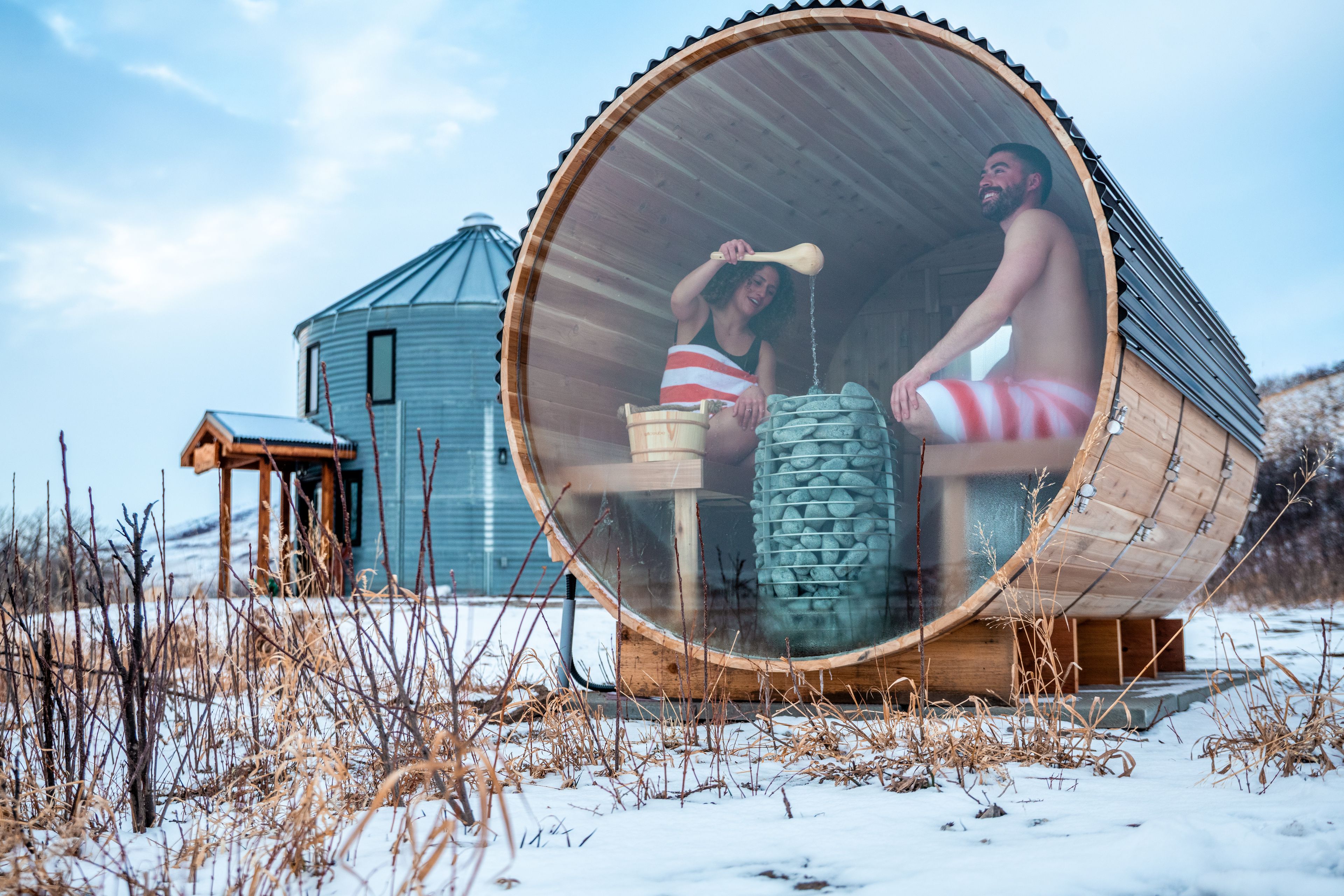 A couple sits in the window of a barrel sauna on a snowy day with their grain silo cabin nearby.