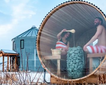 A couple sits in the window of a barrel sauna on a snowy day with their grain silo cabin nearby.