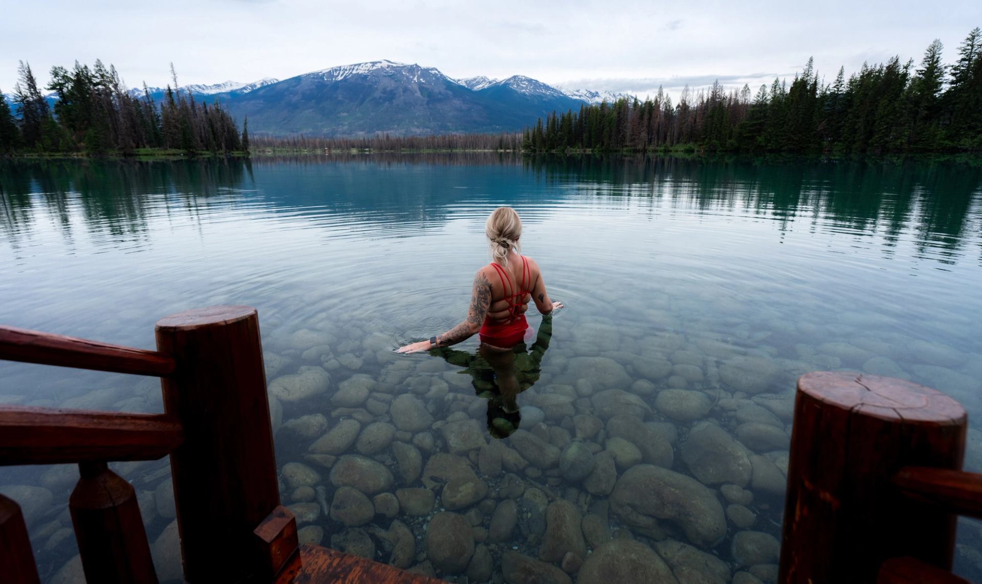 A woman stands in a clear lake during a cold plunge with a mountain and forest in the distance.
