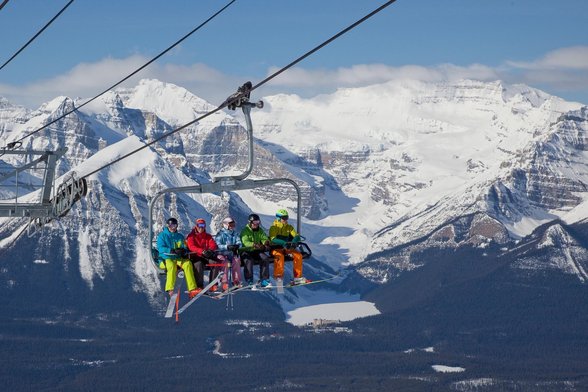 A mix of five skiers and snowboarders riding up a chairlift with mountains in the background.