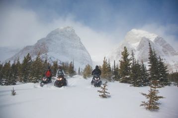 Three snowmobilers riding through snow with a mountain view in the background.