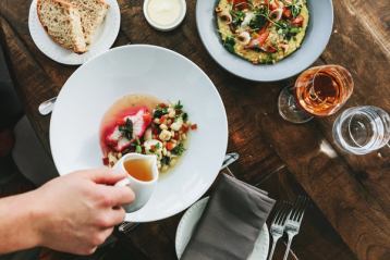 A hand pouring some broth overtop of a colourfully plated dish against a wood grain table