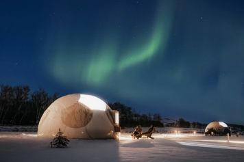 Two people sit outside of a dome accommodation while watching the northern lights. 