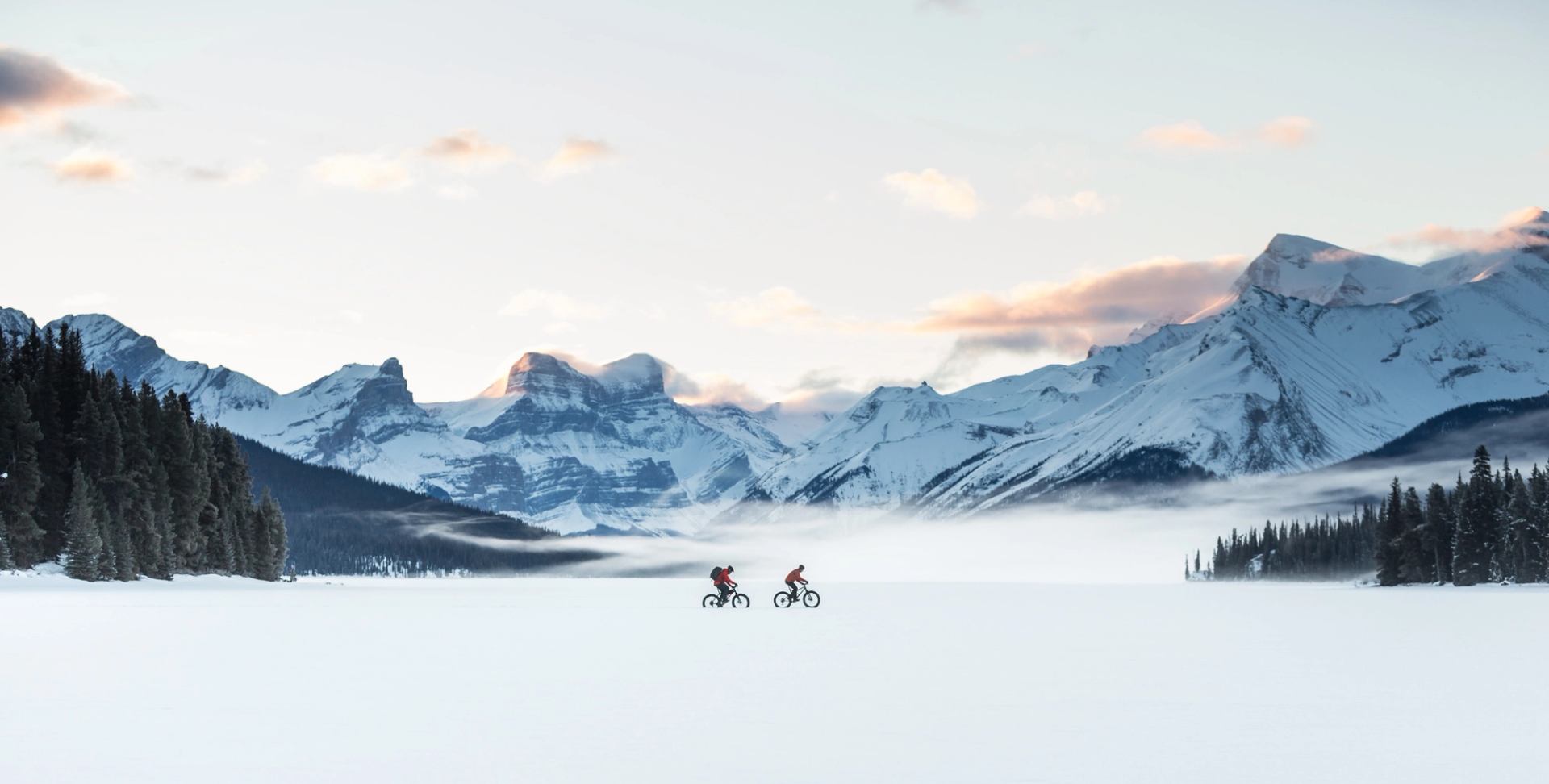 Wide shot of two people fatbiking over a frozen Maligne Lake, mountain range in the background, in Jasper National Park