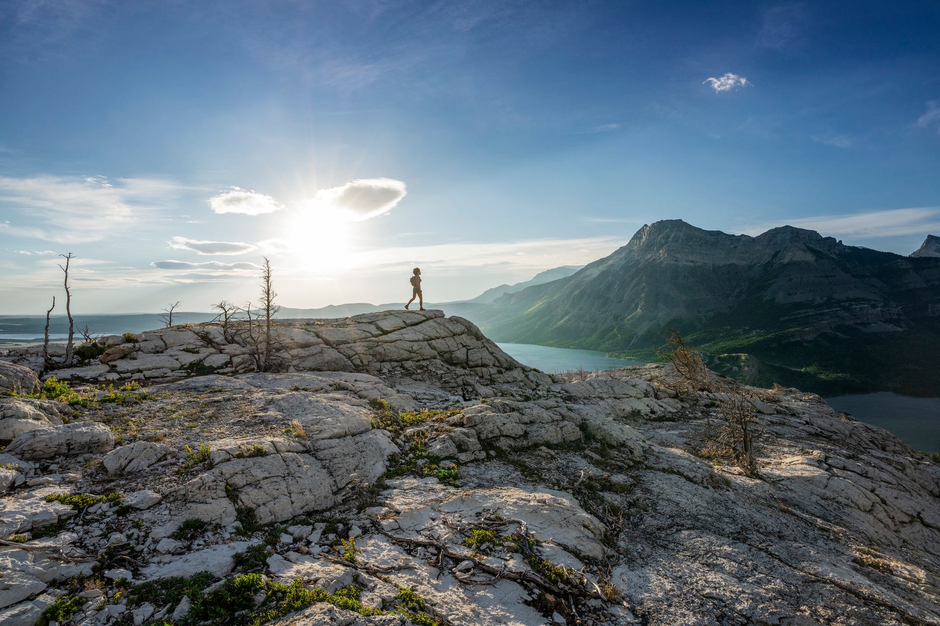 A hiker stands at the tip of a rock looking out over a lake and mountains.