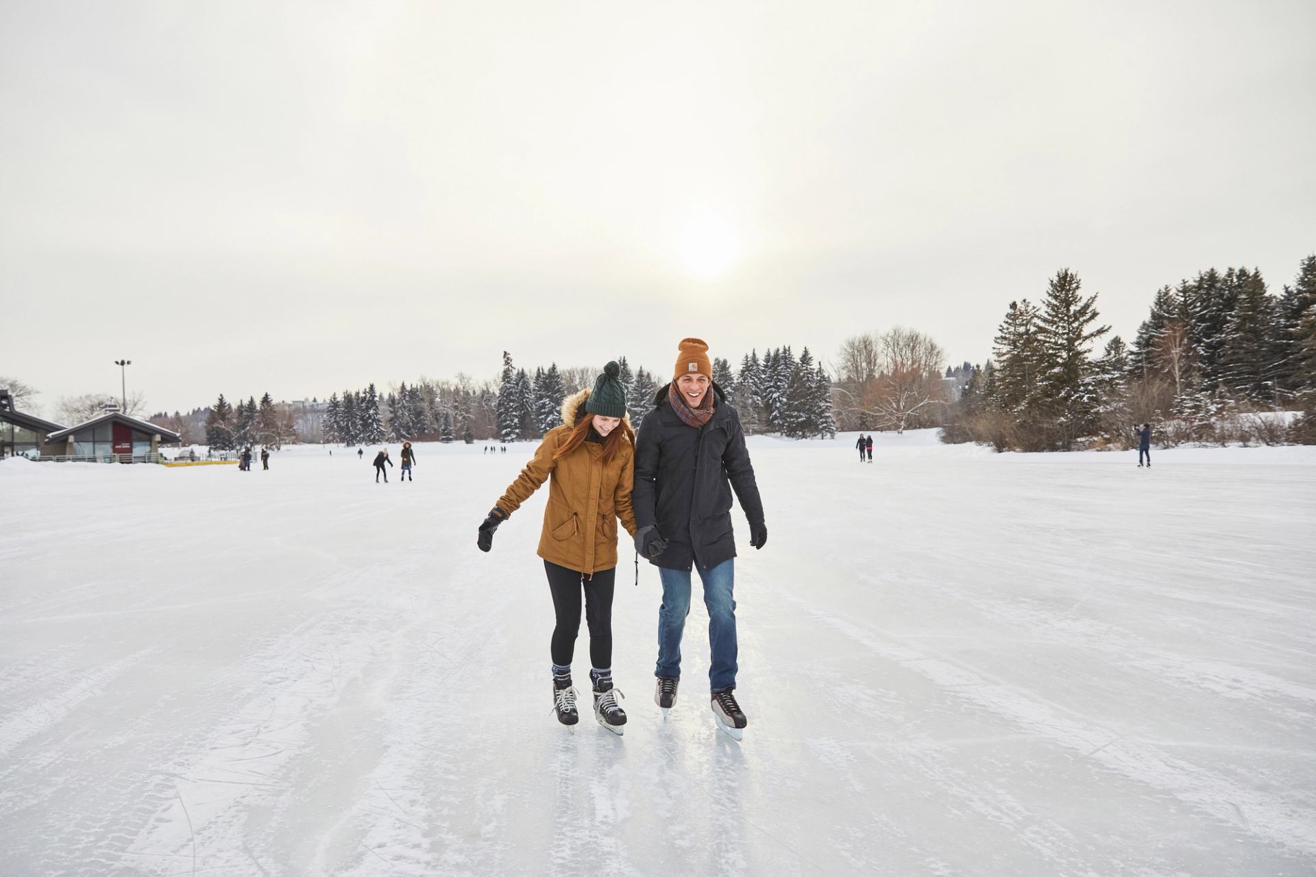 Two people skating on a rink with other skaters and trees in the background.