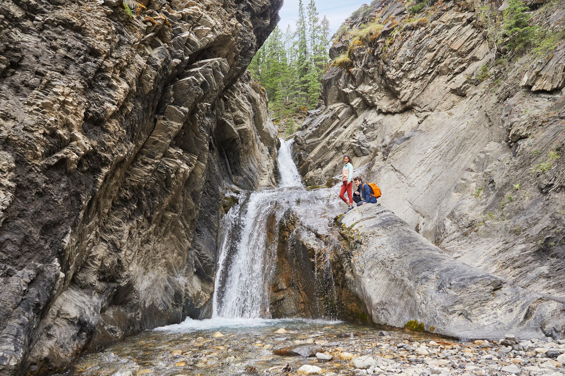 A man and woman stand on a big boulder beside a waterfall.