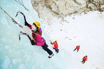 Two people ice climbing a frozen waterfall with two belayers below.