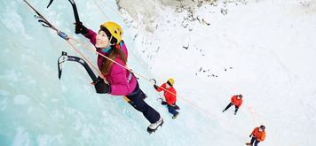 Two people ice climbing a frozen waterfall with two belayers below.