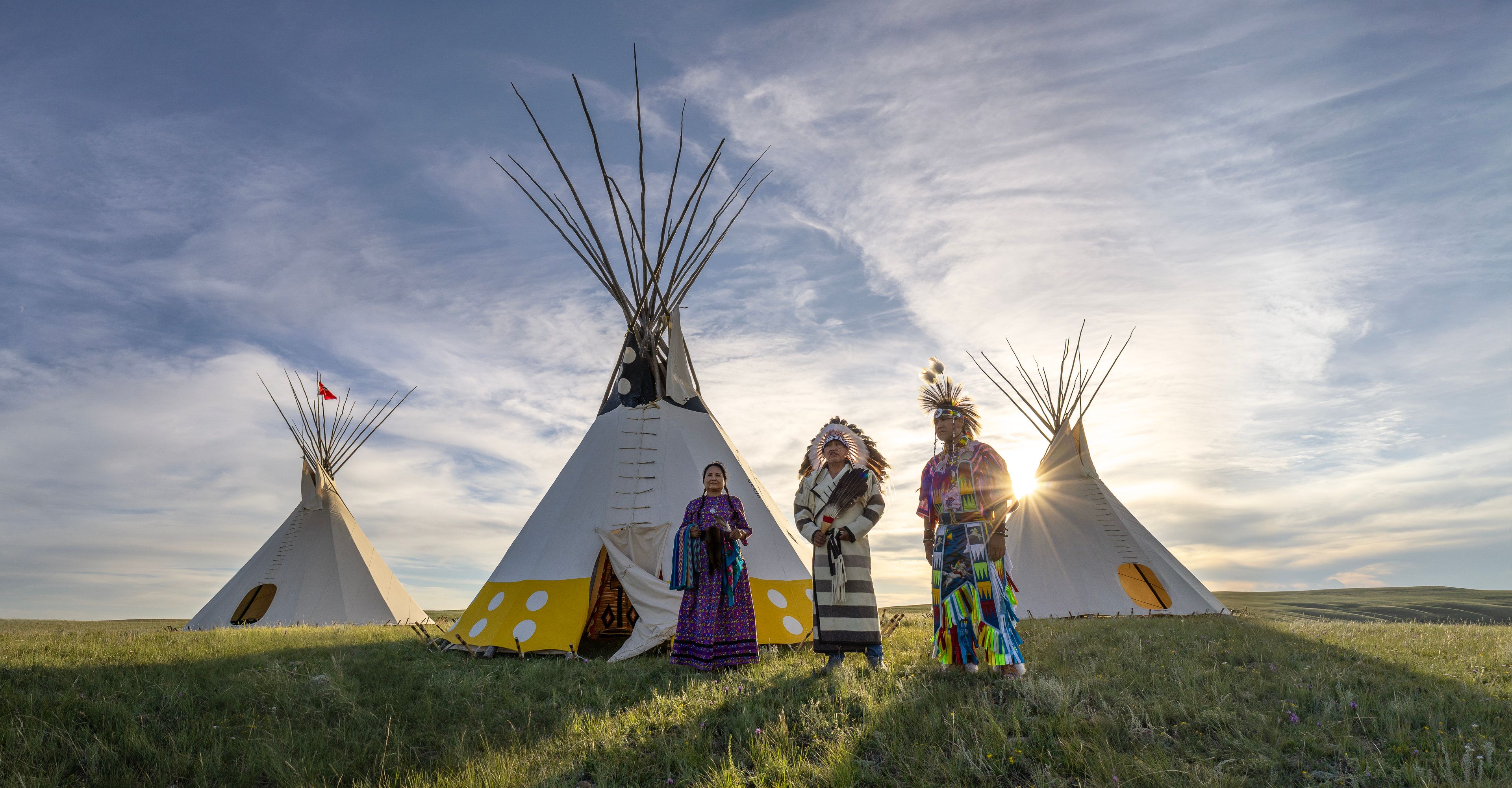 Three people stand in front of Tipis wearing traditional Piikani attire