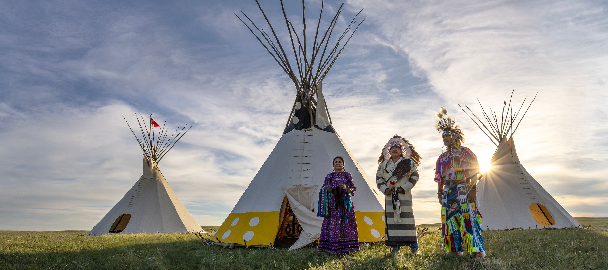 Three people stand in front of Tipis wearing traditional Piikani attire