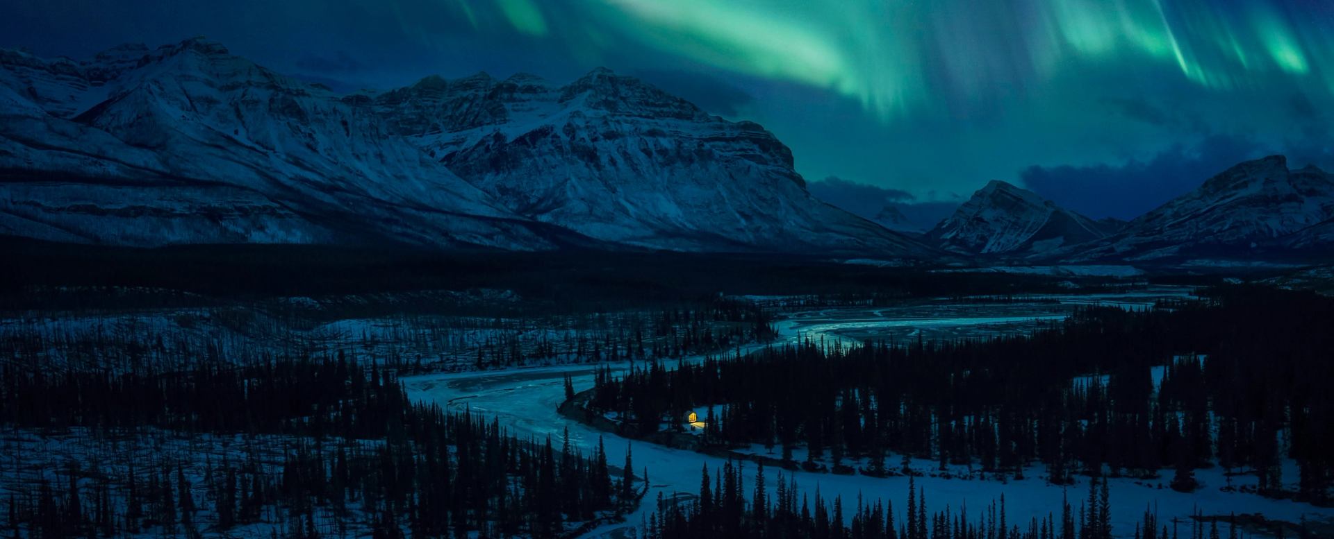 An aerial view of a cabin on the bank of Cline River with the northern lights illuminating the snow-covered Canadian Rockies.