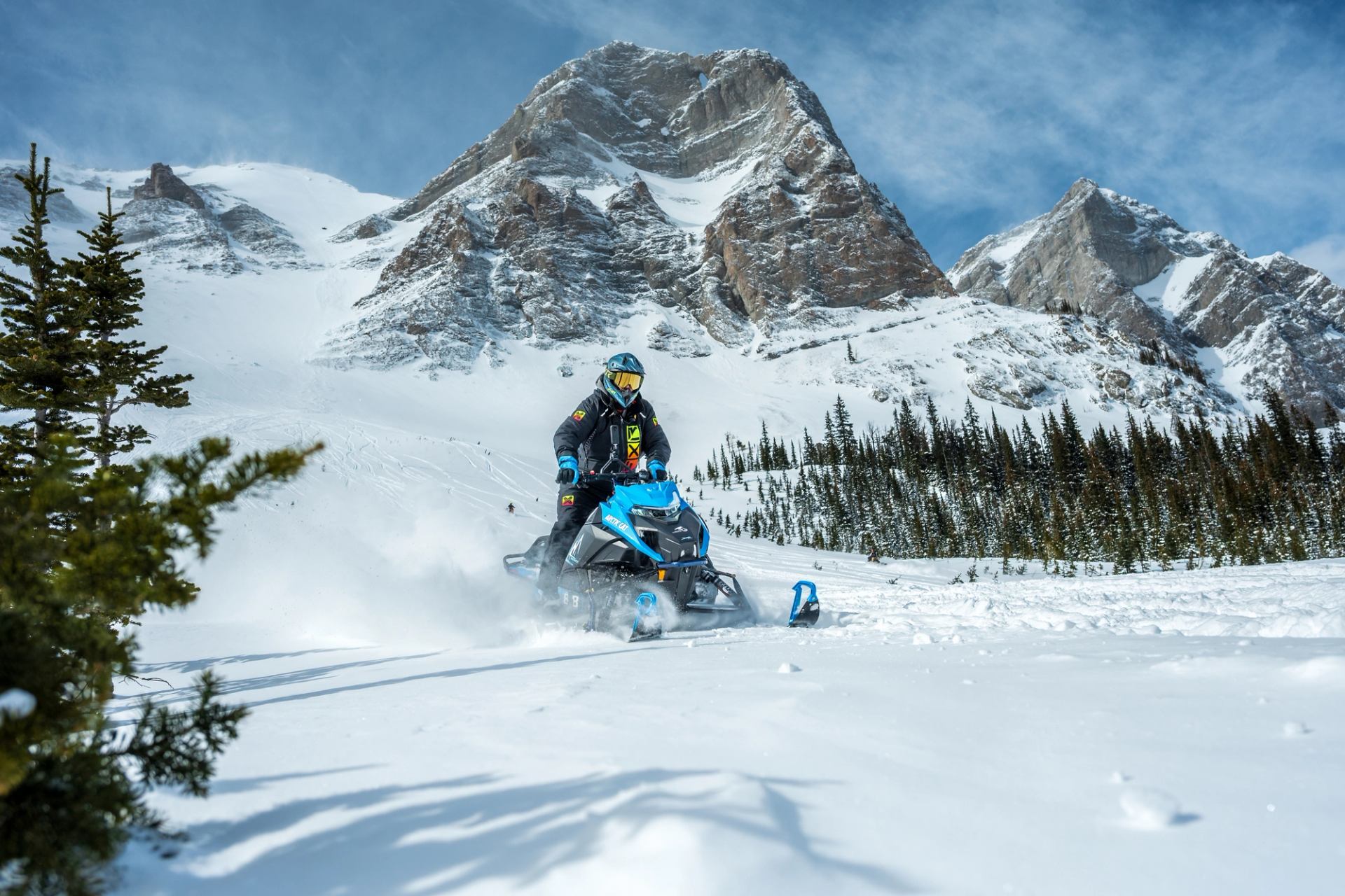 Snowmobiler rides through powder with majestic mountains in background.