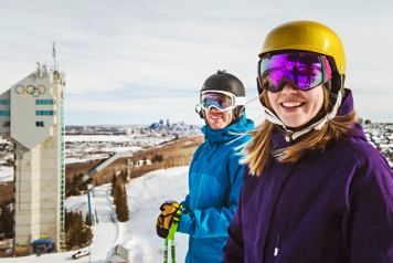 Two skiers at the top of a run at WinSport with the Olympic ski jump tower in the distance.